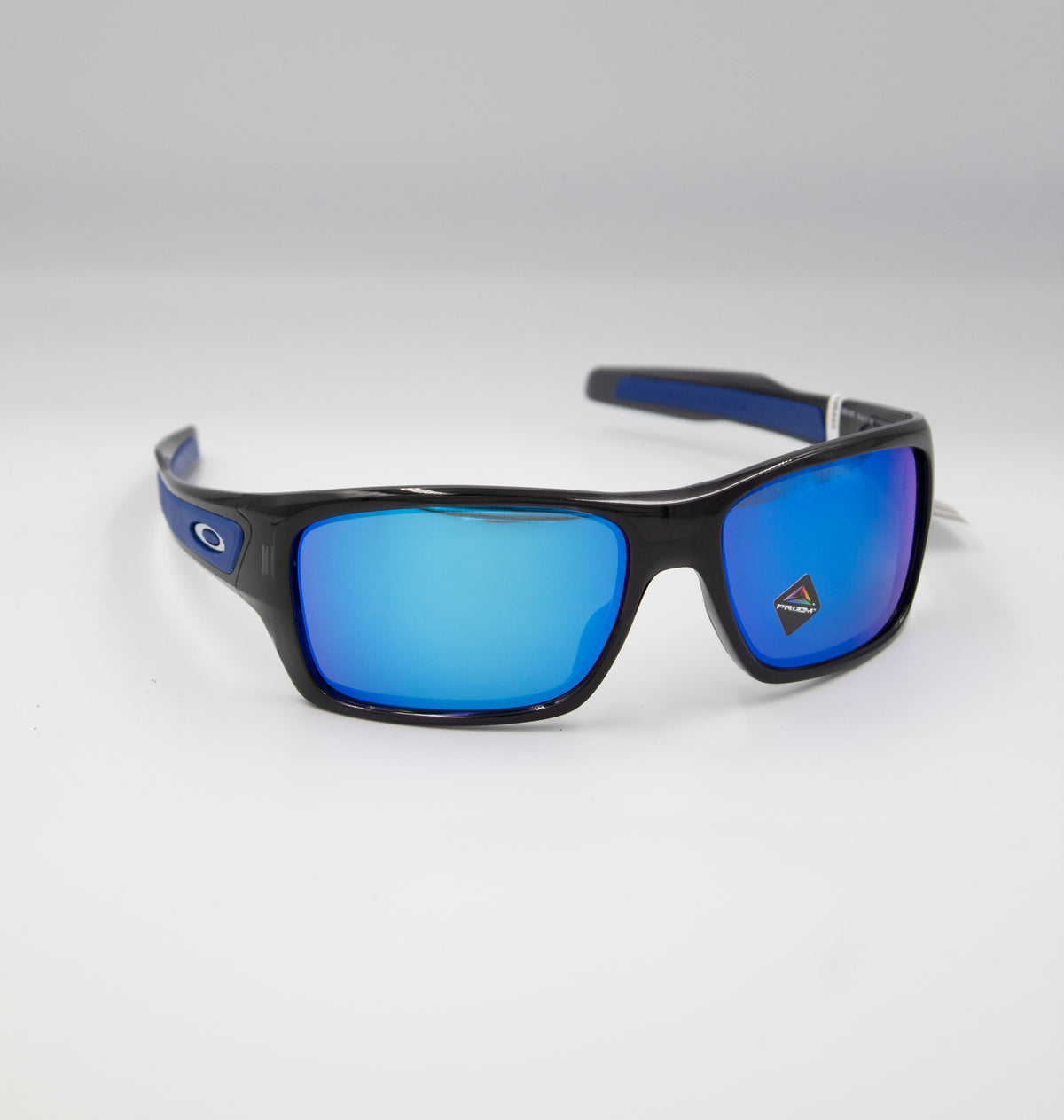The Oakley Turbine sunglasses boast a sleek black frame with vibrant blue-tinted lenses. Positioned against a plain white background, they highlight subtle blue temple details and a discreet logo on the left temple.