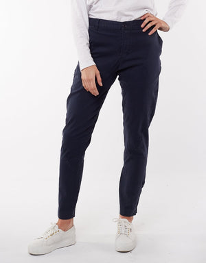 A person in navy blue Foxwood Liberty Pants and a white long-sleeve shirt is standing with their right hand placed on their hip. They are also wearing white sneakers. The comfortable pants have a relaxed fit with a tapered leg, perfect for casual wear. The background is plain white.
