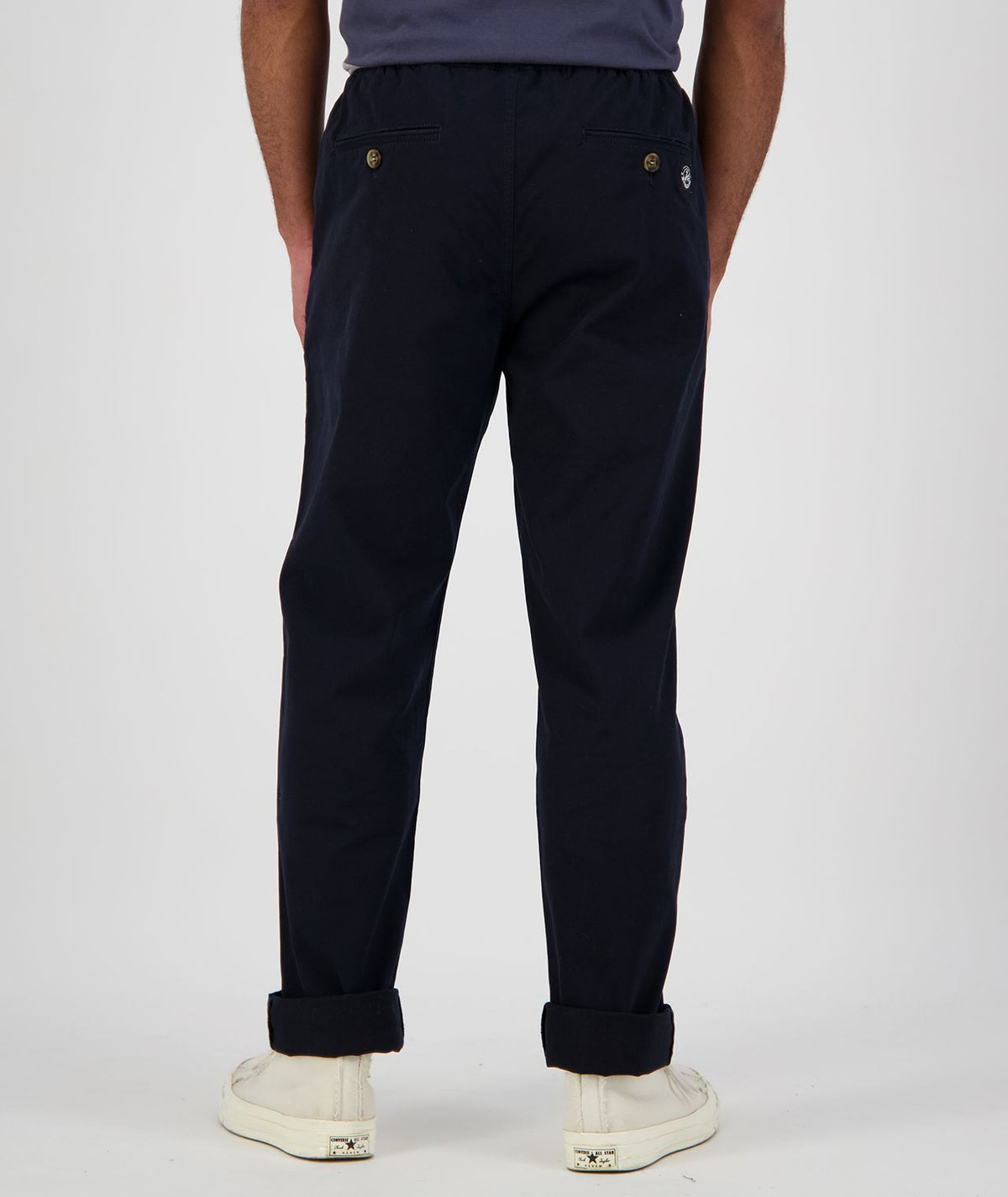 A person is standing against a plain background, facing away. They are wearing black cuffed Swanndri Karaka Pants, a dark shirt, and white sneakers. The cotton pants feature buttons and pockets at the back. Only the lower half of the person's body is visible.