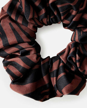 A close-up of the Rip Curl Sun Tribe Scrunchie reveals a black and dark brown zebra pattern on its knit fabric. The scrunchie, arranged in a circular shape, highlights its texture and design against a plain white backdrop.