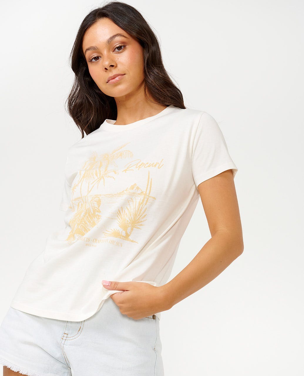 A woman with long dark hair stands against a plain background, showcasing her stylish outfit: the Rip Curl Buena Vista Standard Tee, a soft cotton jersey t-shirt in white with a yellow graphic design, paired with light denim shorts.