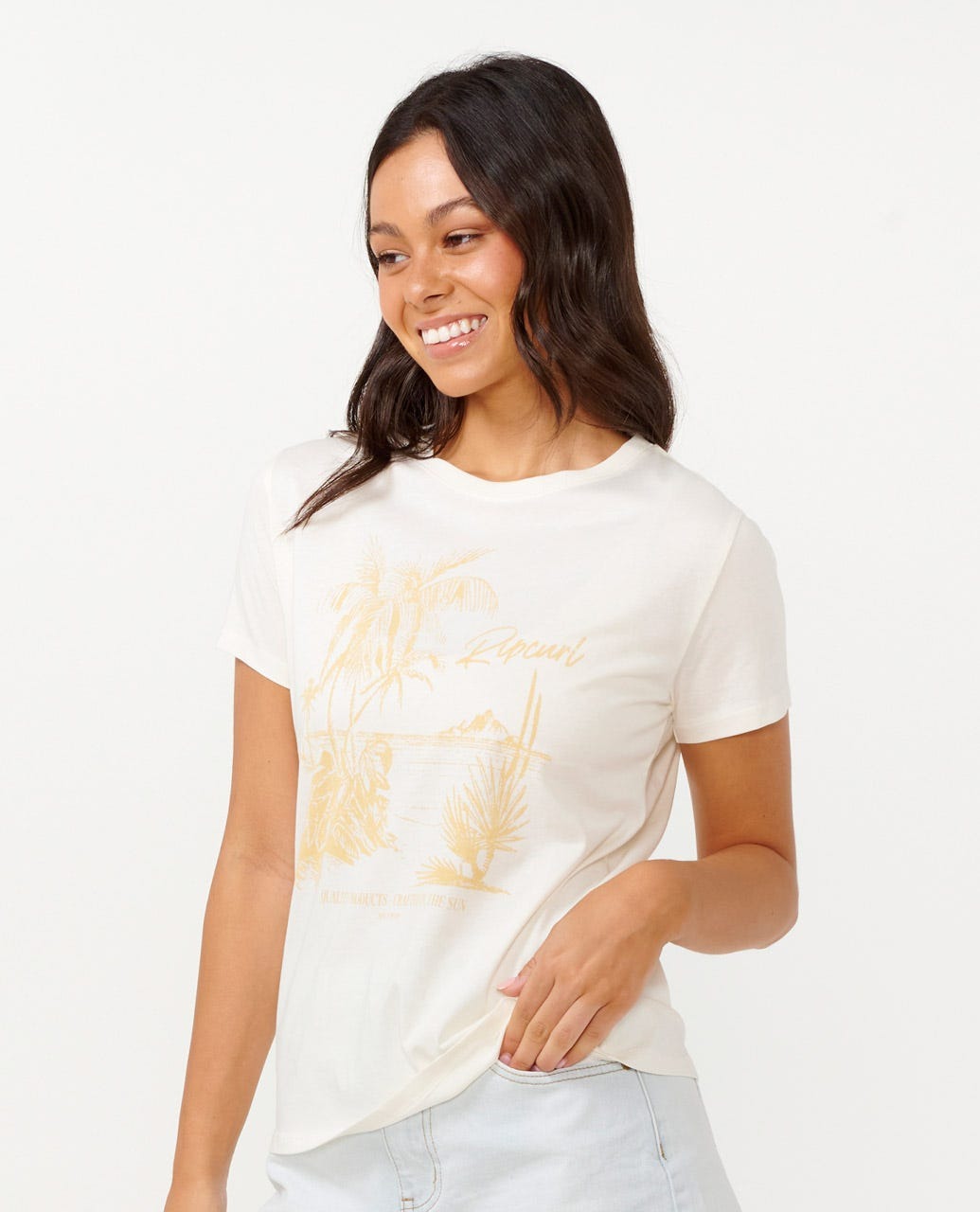 A person with long dark hair smiles while wearing a Rip Curl Buena Vista Standard Tee. Made from cotton jersey in a standard fit, it showcases a tropical design. They are standing against a plain background, holding one hand near their hip and looking slightly to the side.