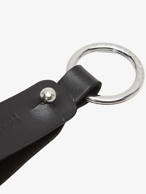 This close-up features the RM Williams Pioneer Key Fob, a black double butt leather keychain with nickel hardware. The leather is secured by a sturdy metal rivet, and the silver ring, functioning as a key fob, displays engraved text around its outer edge.