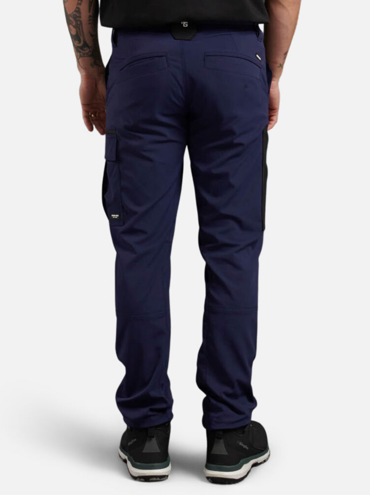 A person stands against a plain white background wearing navy King Gee Trademark Cargo Pants with black patches, black sneakers, and a black shirt. The ripstop fabric pants feature multiple pockets, and they have a tattoo on their left arm.