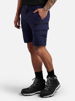 A person in King Gee Trademark Cargo Shorts, navy in color and made from stretch ripstop fabric, stands with their left hand in the side zipper and flap pocket. They're wearing black hiking boots with gray laces and showcase a tattoo on their left arm against a plain background.