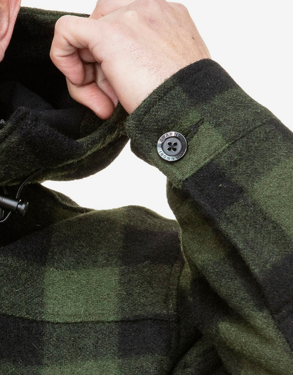 A person adjusts the hood of a Swanndri Hudson Hoody, a warm green and black plaid jacket made from New Zealand wool. It features button detail on the sleeve near the wrist, with the hand partially visible and fingers slightly curled.