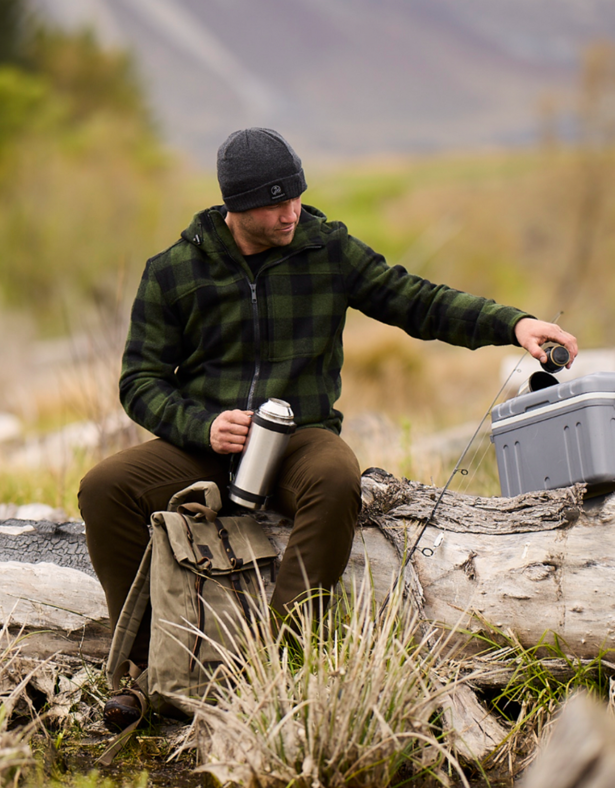 A man in a green Swanndri Hudson Hoody and black beanie sits on a fallen log in a grassy area, holding a thermos. He reaches for the cooler beside him, while his backpack rests nearby. The blurred trees and hills backdrop completes this cozy scene amidst nature.