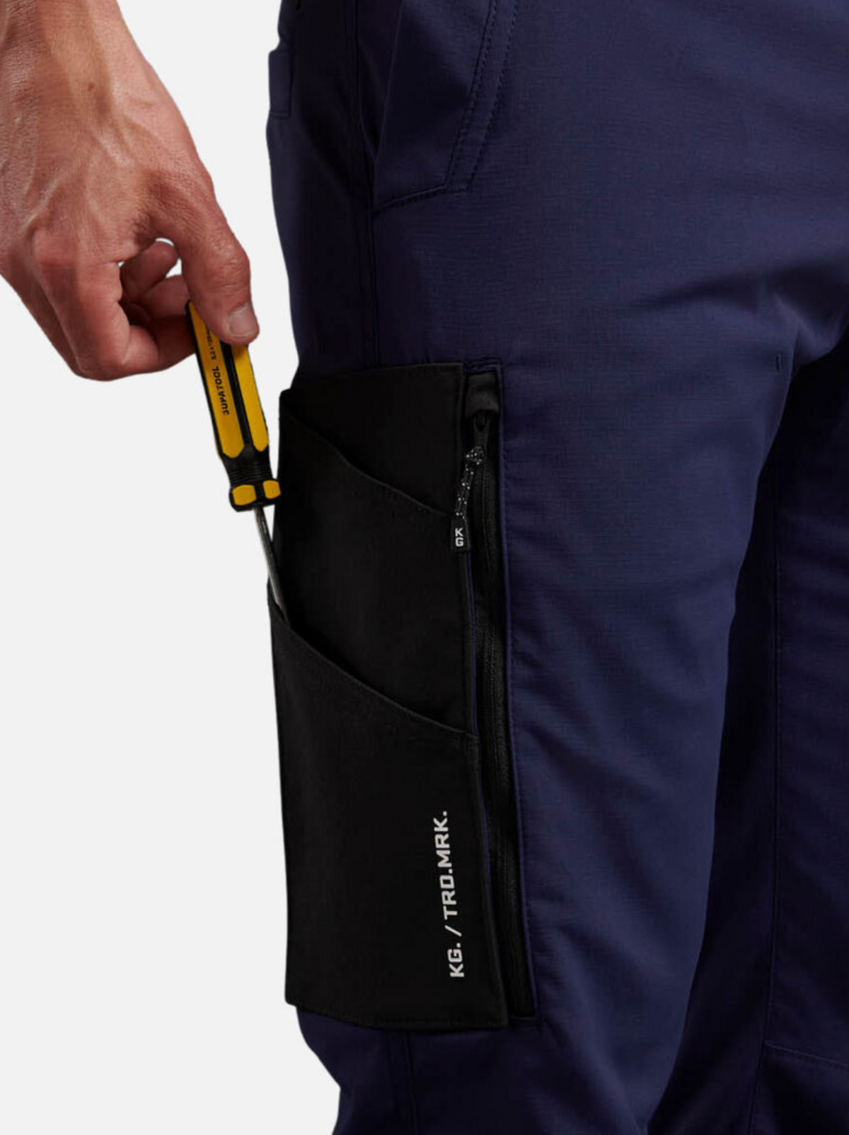 A person slips a yellow-handled tool into the ripstop fabric pocket of their navy blue King Gee Trademark Cargo Pant, featuring a black, zippered compartment with "TIDAL/ARK." embroidery for durable style.