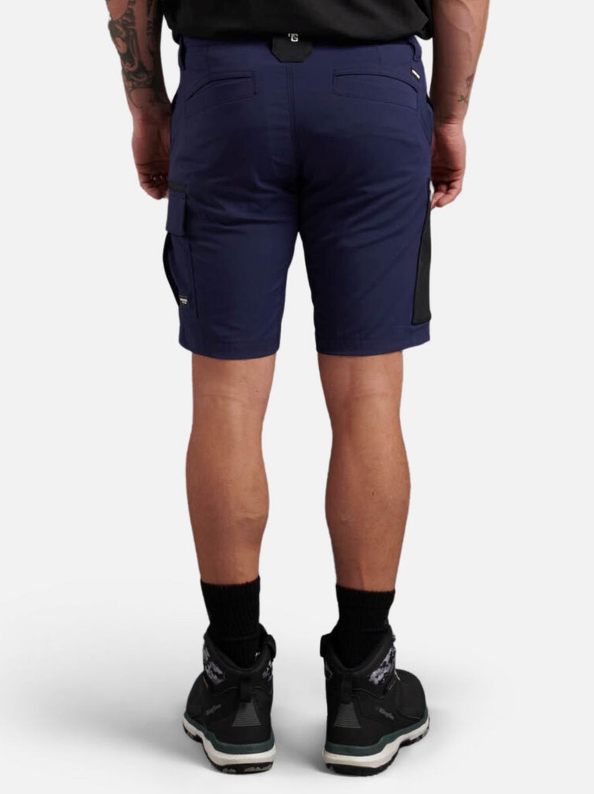 Rear view of a person wearing King Gee Trademark Cargo Shorts in navy blue with black pockets, paired with black sneakers and socks. The individual also sports a shirt and displays tattoos on their arm, all set against a plain white background.