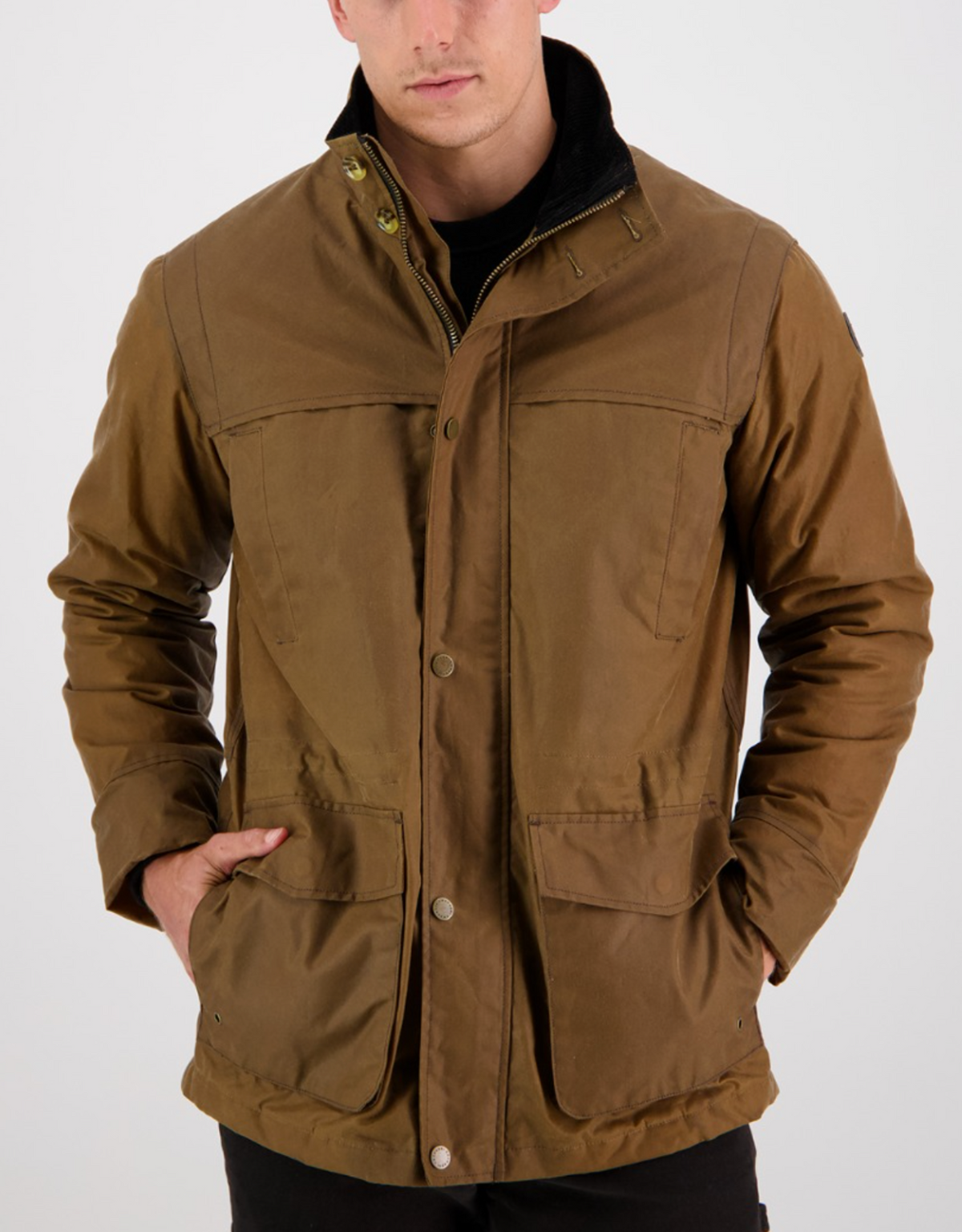 A person is seen wearing the Swanndri Evans Flat Oilskin Jacket, distinguished by its brown color, high collar, and button closure. The jacket has large front pockets where the person's hands rest casually. The background is simple and white.