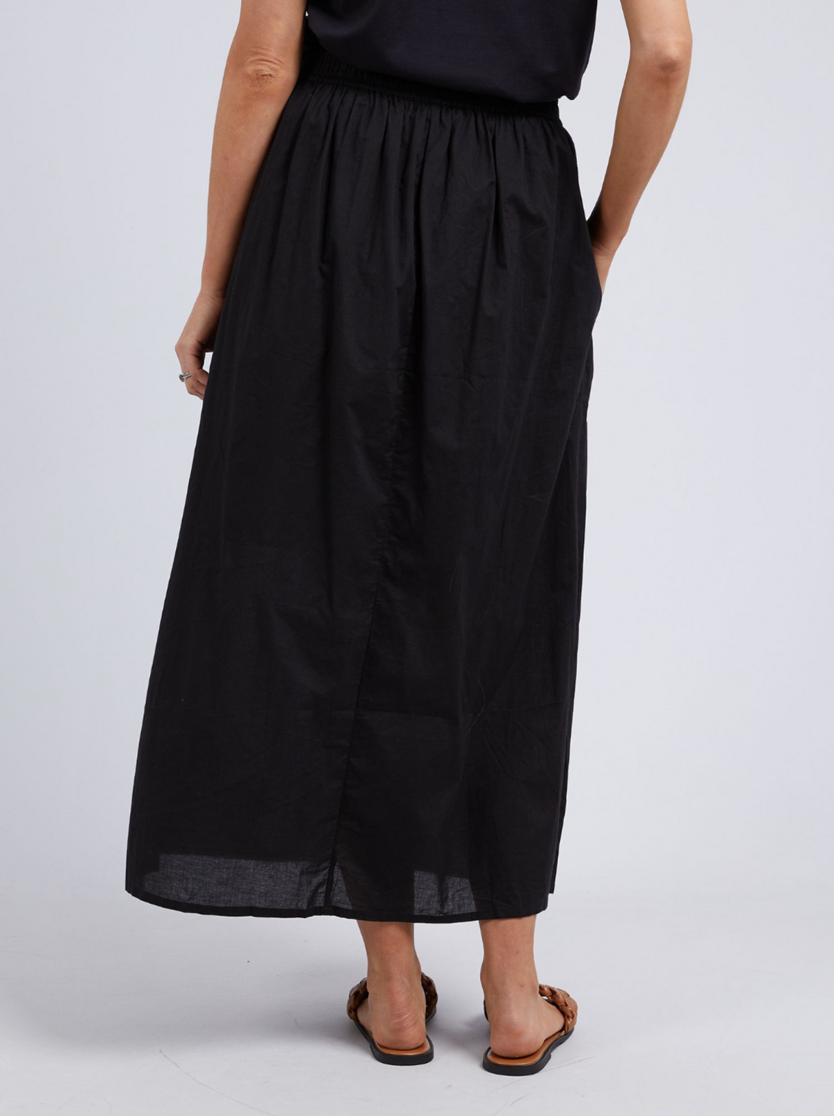 A person wearing the Foxwood Charli Skirt—a long black maxi with an elastic waist—paired with a black top and brown flat sandals stands with their back to the camera against a plain light gray background.