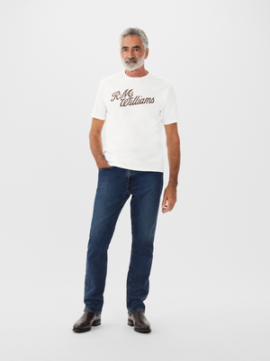 A man with gray hair and a beard wears a regular fit, white cotton RM Williams R.M.W Script T-Shirt, paired with blue jeans and brown boots. His left hand rests in his pocket against a plain white background.