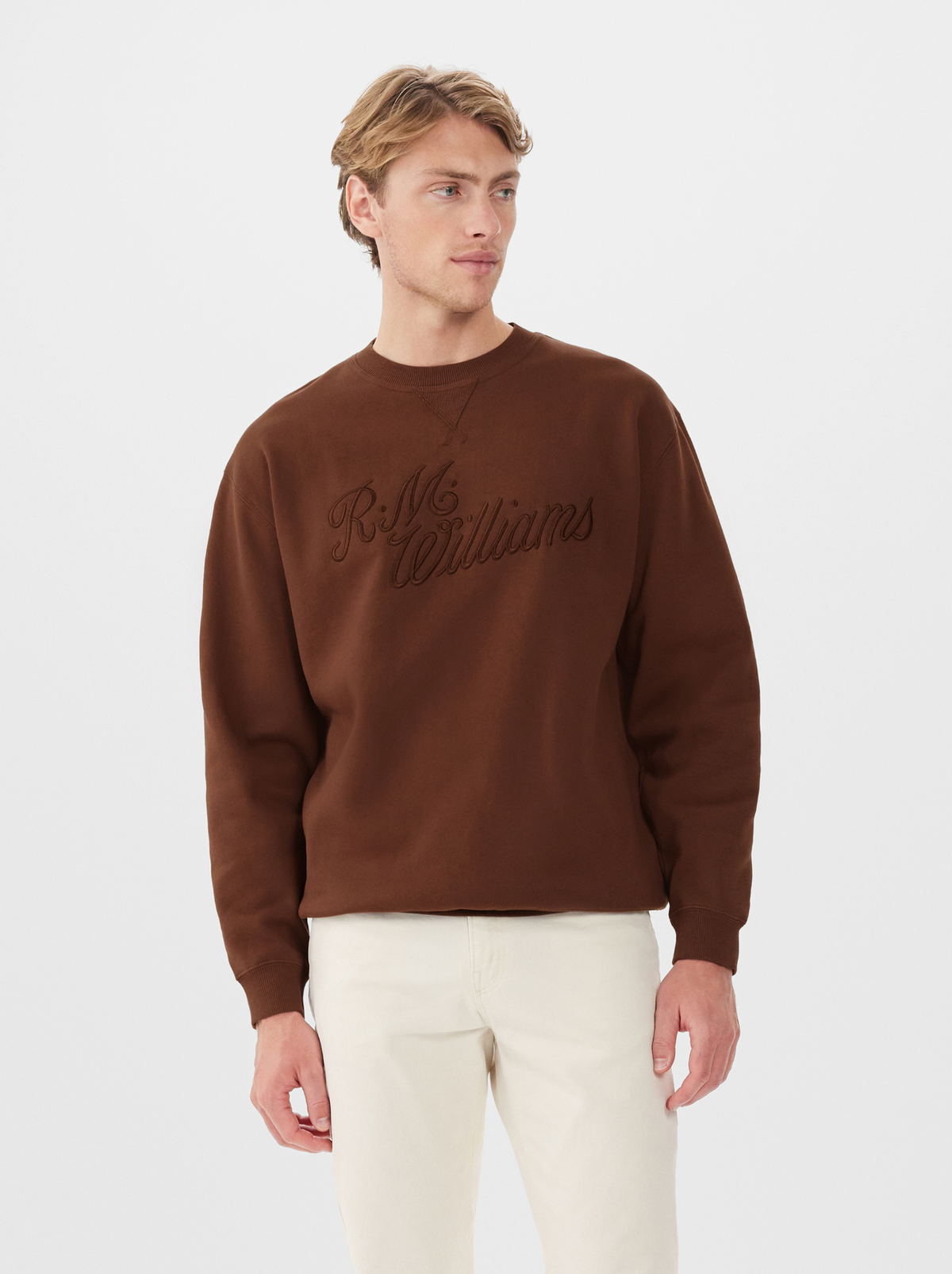 A man wearing a brown RM Williams R.M.W Script Crew Neck jumper and off-white pants stands against a plain white background, looking slightly to his left.