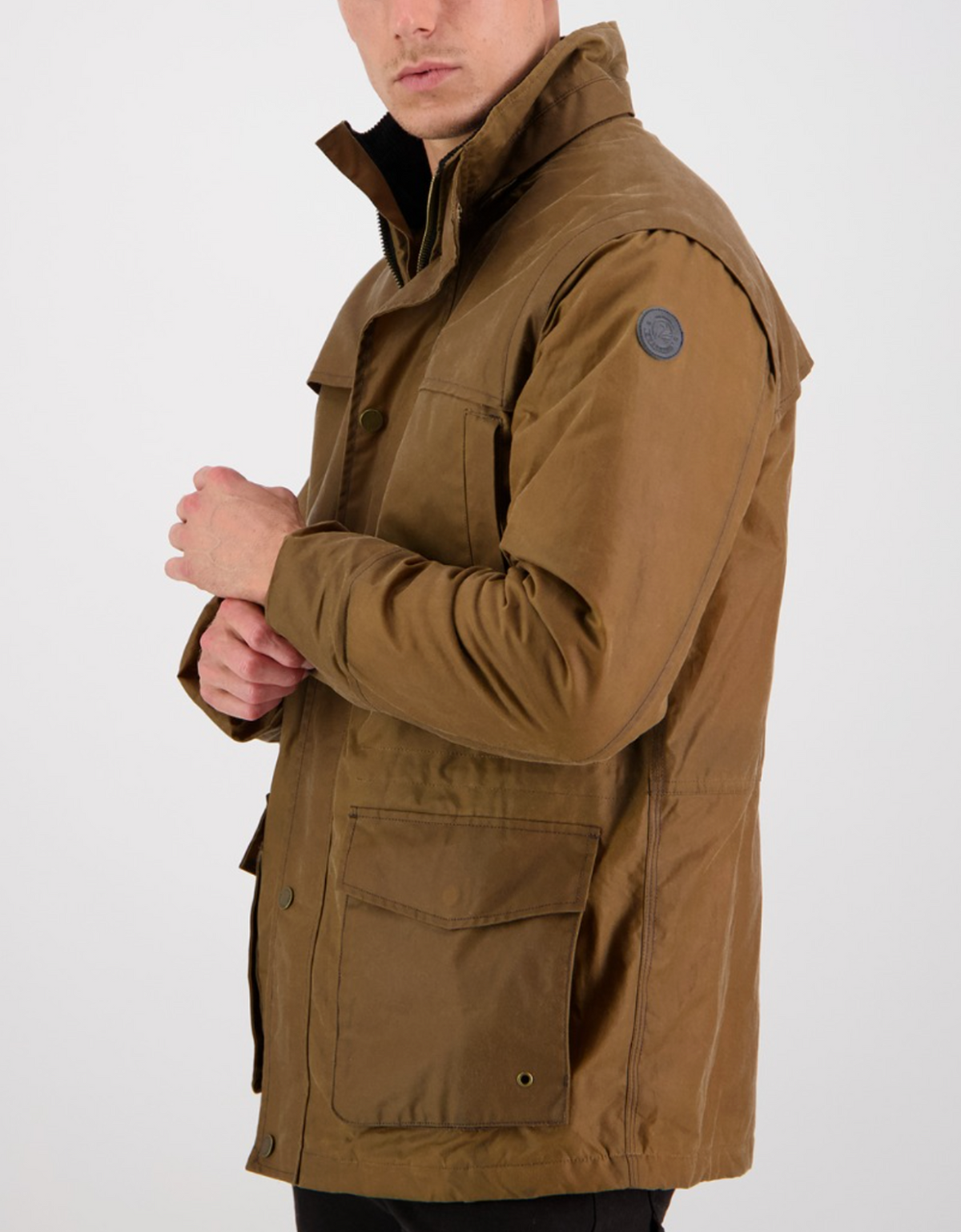 A person in a Swanndri Evans Flat Oilskin Jacket, featuring a high collar, buttoned front, large pockets, and a circular sleeve patch. They are posing sideways with arms partially crossed against a light background.