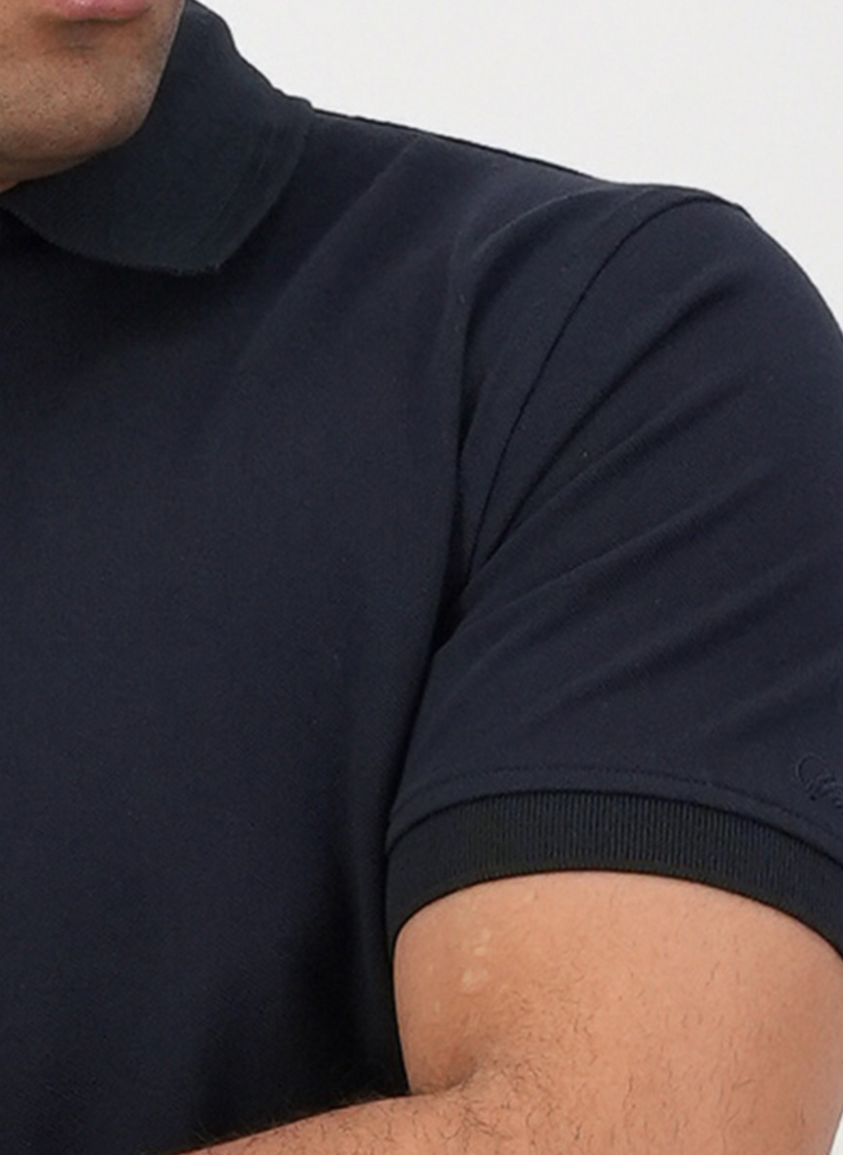 A close-up shows a person wearing the Swanndri Highgrove Polo in dark navy, made from 100% combed cotton, with ribbed collar and sleeve trim. Part of their face, shoulder, and crossed arm are visible.