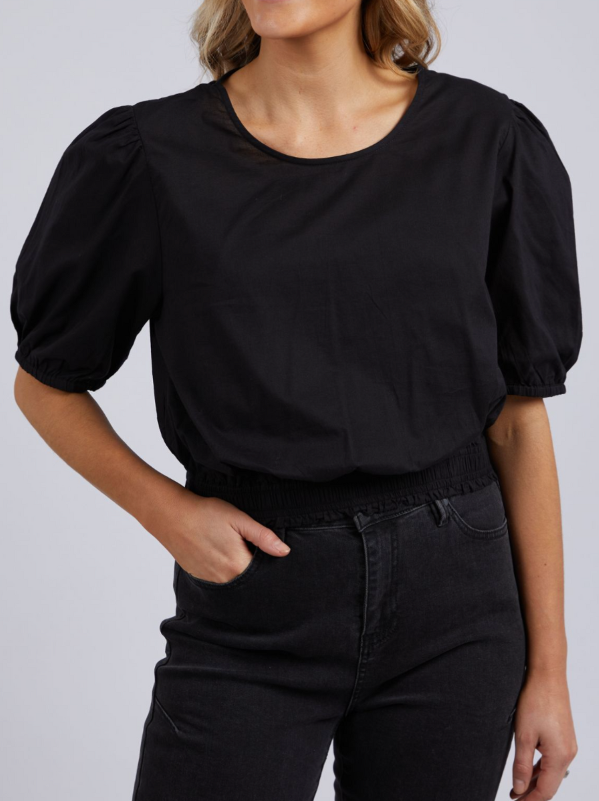 A person wears the Foxwood Charli Top, a black cropped fit blouse with puff sleeves, paired with high-waisted black jeans. One hand is in their pocket against a plain gray background.