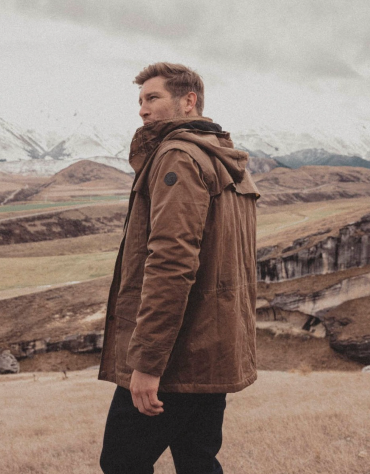 A person in a Swanndri Evans Flat Oilskin Jacket stands before grassy hills and snow-capped mountains under a cloudy sky.