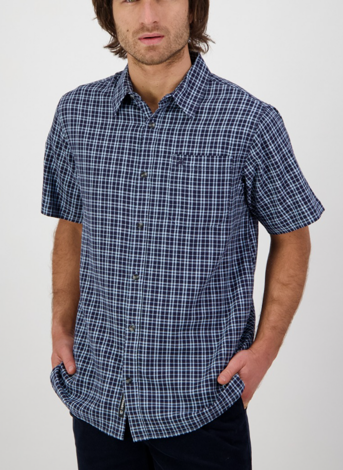 Someone in a relaxed fit Swanndri Russell Shirt, featuring 100% cotton with short sleeves and a dark blue and white checkered design, stands with hands in pockets against a plain white background.