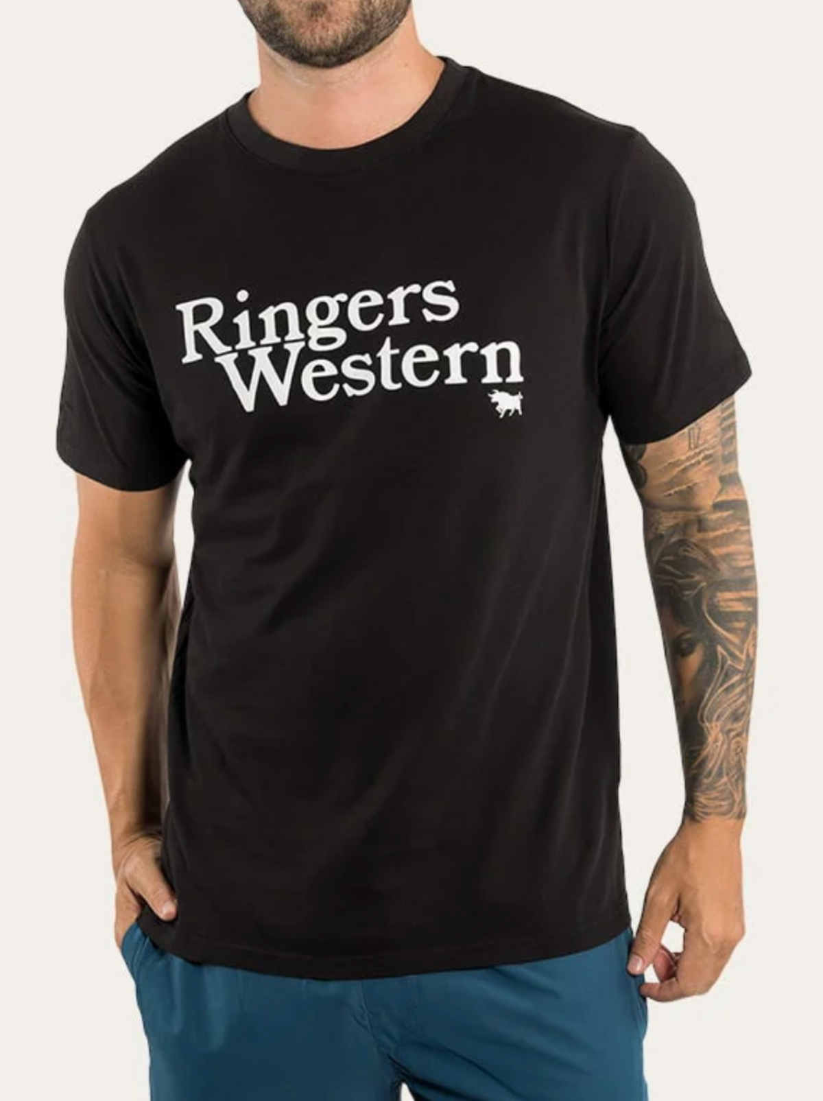 A man wearing the Ringers Western The Lodge Classic Fit T Shirt, made from 100% cotton jersey with white text, and blue shorts. He has a tattoo on his right arm and one hand in his pocket.