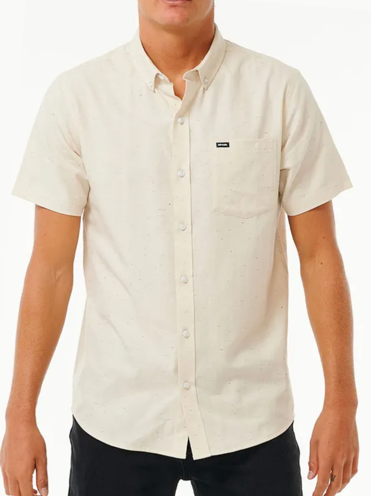 Someone wears the Rip Curl Ourtime Short Sleeve Shirt in cream, featuring a chest pocket and small black label, with black pants. The shirt has a subtle speckled texture and is made from 100% cotton herringbone.