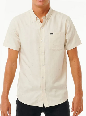 Someone wears the Rip Curl Ourtime Short Sleeve Shirt in cream, featuring a chest pocket and small black label, with black pants. The shirt has a subtle speckled texture and is made from 100% cotton herringbone.