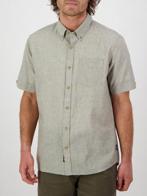 A man stands against a white background wearing the Swanndri Lowell v2 Short Sleeve Shirt, light gray with an embroidered swan logo on the chest pocket. Made from a linen-cotton blend, it’s a classic fit. His relaxed arms frame his wavy brown hair partially covering his face.
