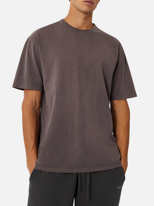 A man wears an oversized, washed dark brown Industrie The Del Sur Tee (100% cotton, short sleeves) with black drawstring pants, standing against a plain white background. His face is partially out of frame.