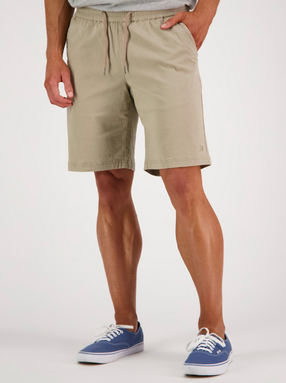 Wearing Swanndri Clevedon Stretch Canvas Shorts with an elastic waist and blue canvas sneakers, a person stands against a plain white background, showing only their lower torso and legs.