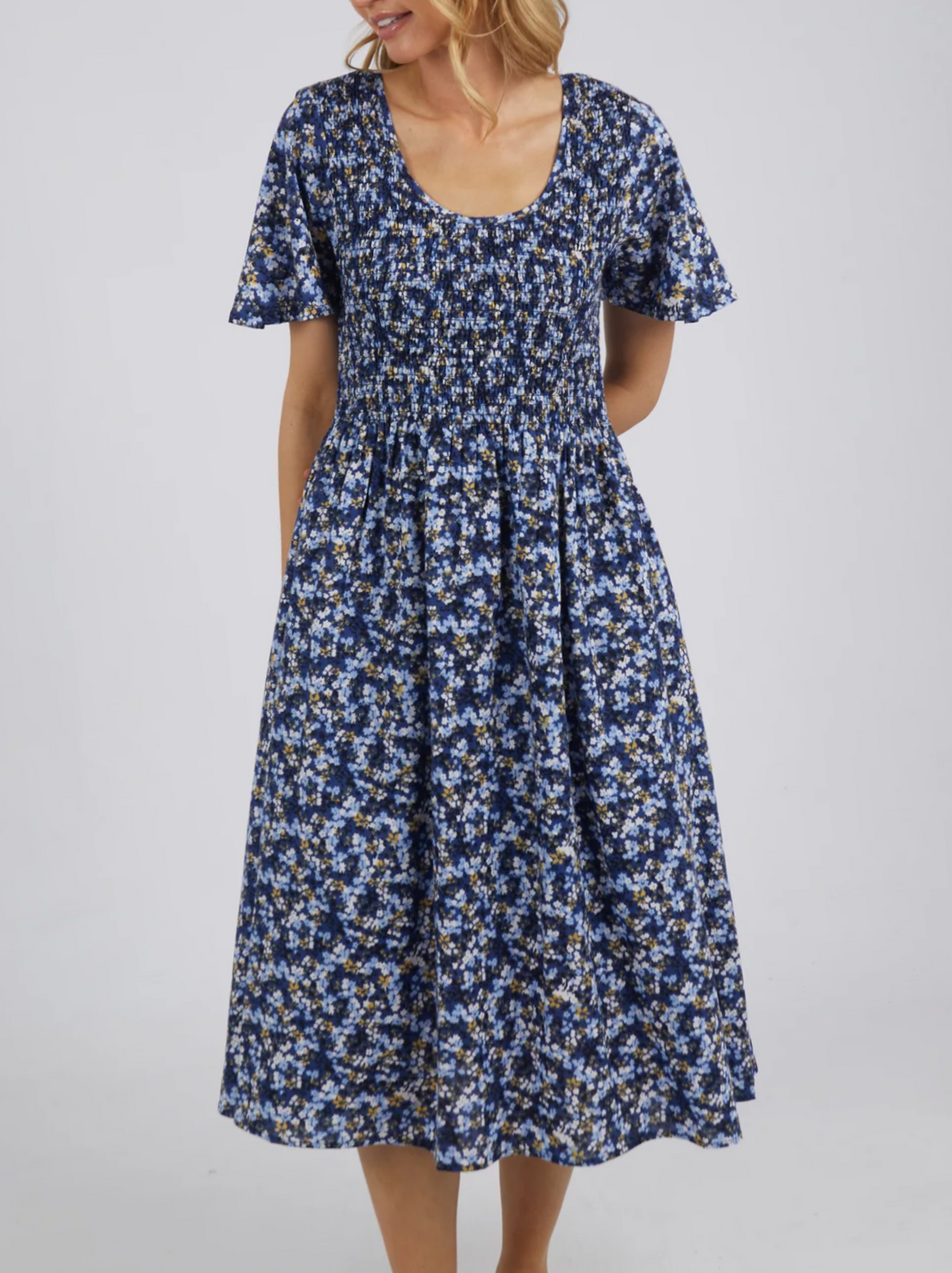A woman wears the Foxwood Lulu Dress in sapphire ditsy print, made from cotton poplin with a smocked bodice and flared skirt. She stands against a plain light background.