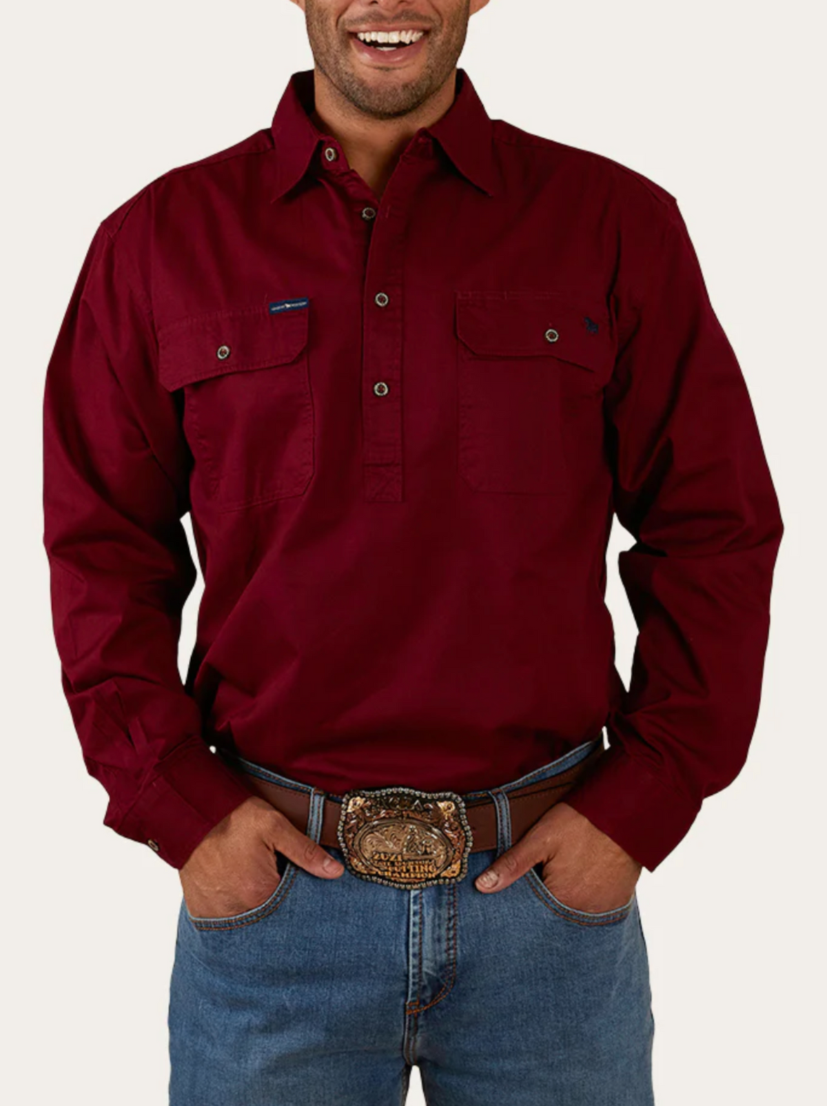 A man is smiling with his hands in his pockets, wearing a maroon Ringers Western King River Half Button Shirt with two chest pockets, blue jeans, and a large decorative belt buckle.