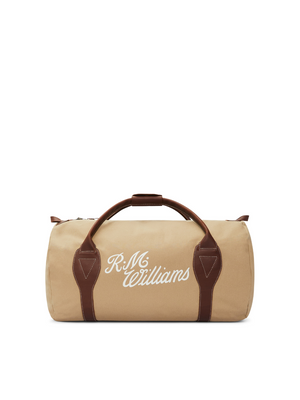 The RM Williams Sorrento Ute Bag is a beige cylindrical duffel featuring brown leather straps and handles, embellished with white embroidered "R.M. Williams" text on the side. Made from water-repellant utility canvas, it includes a top zipper closure for added practicality.