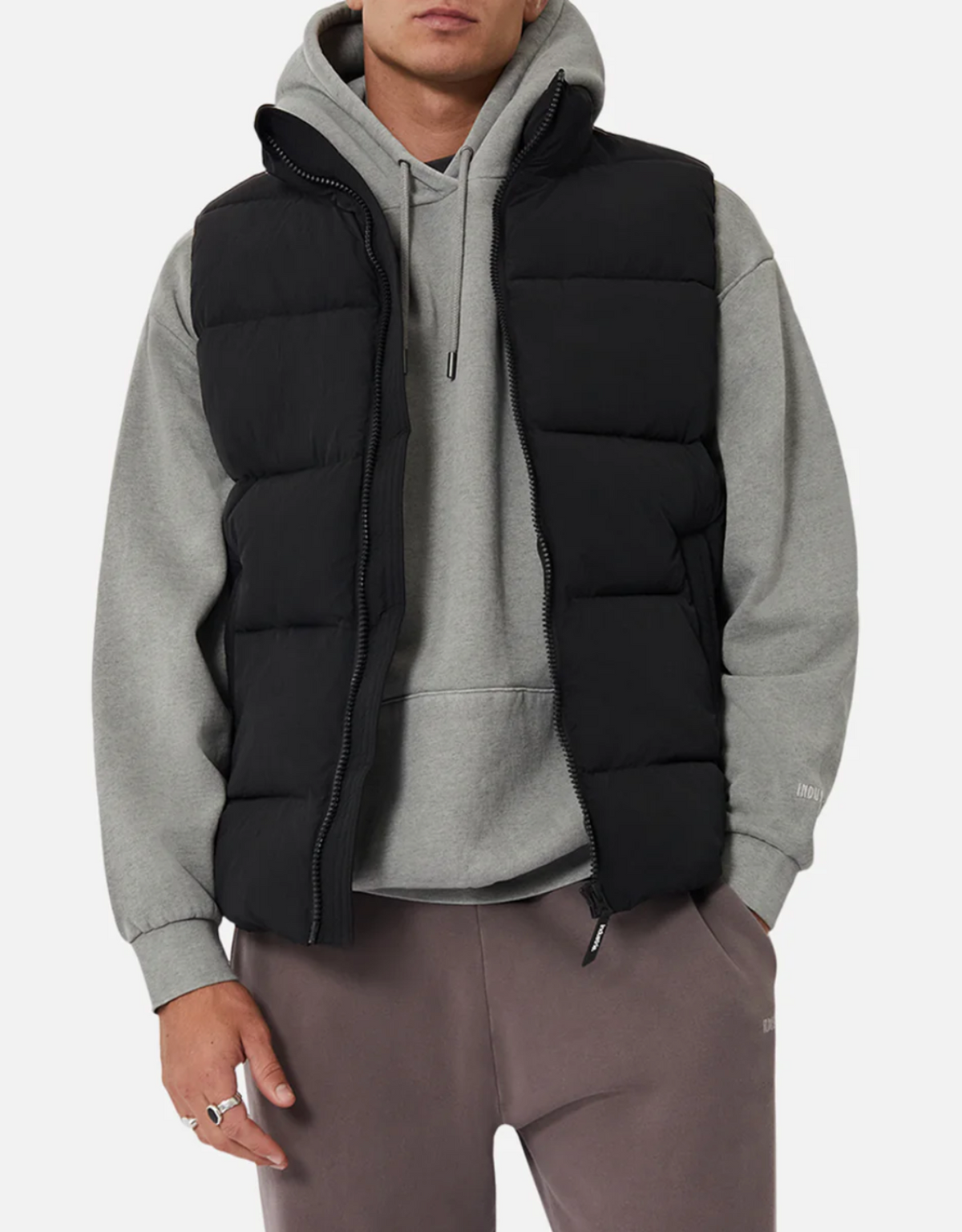 Someone is wearing a gray hoodie layered under an Industrie The New Hanam Puffer Vest with quilted padding, paired with dark gray pants. Their hand is partially visible at their side against a plain white background.