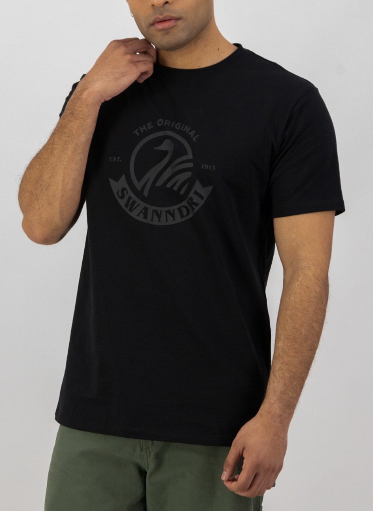 A man in a black Swanndri Original v2 Print T-Shirt, made from organic cotton with a circular emblem and text, stands with arms relaxed in olive green pants against a plain white background, embodying everyday style.