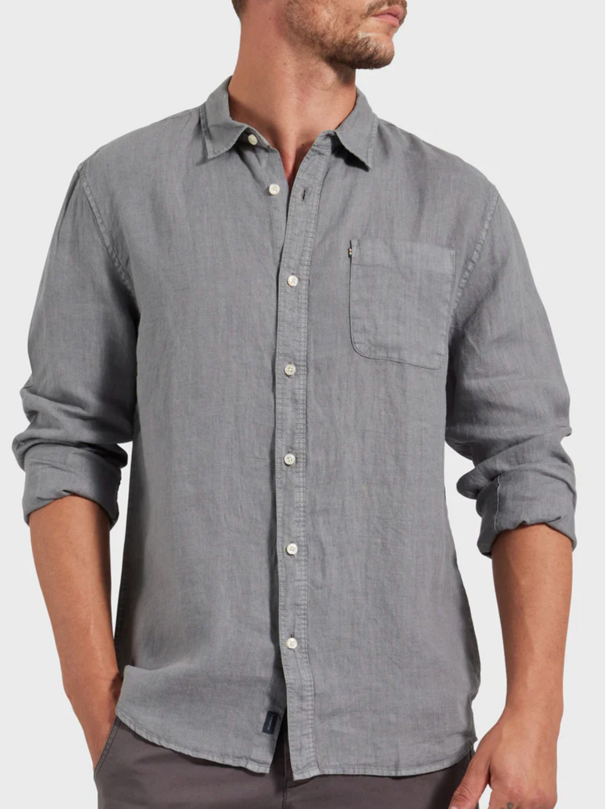 A man stands with hands in his pockets, looking to the side, wearing the Academy Brand Hampton Linen Shirt in stylish light gray, featuring a chest pocket and slightly rolled-up long sleeves.