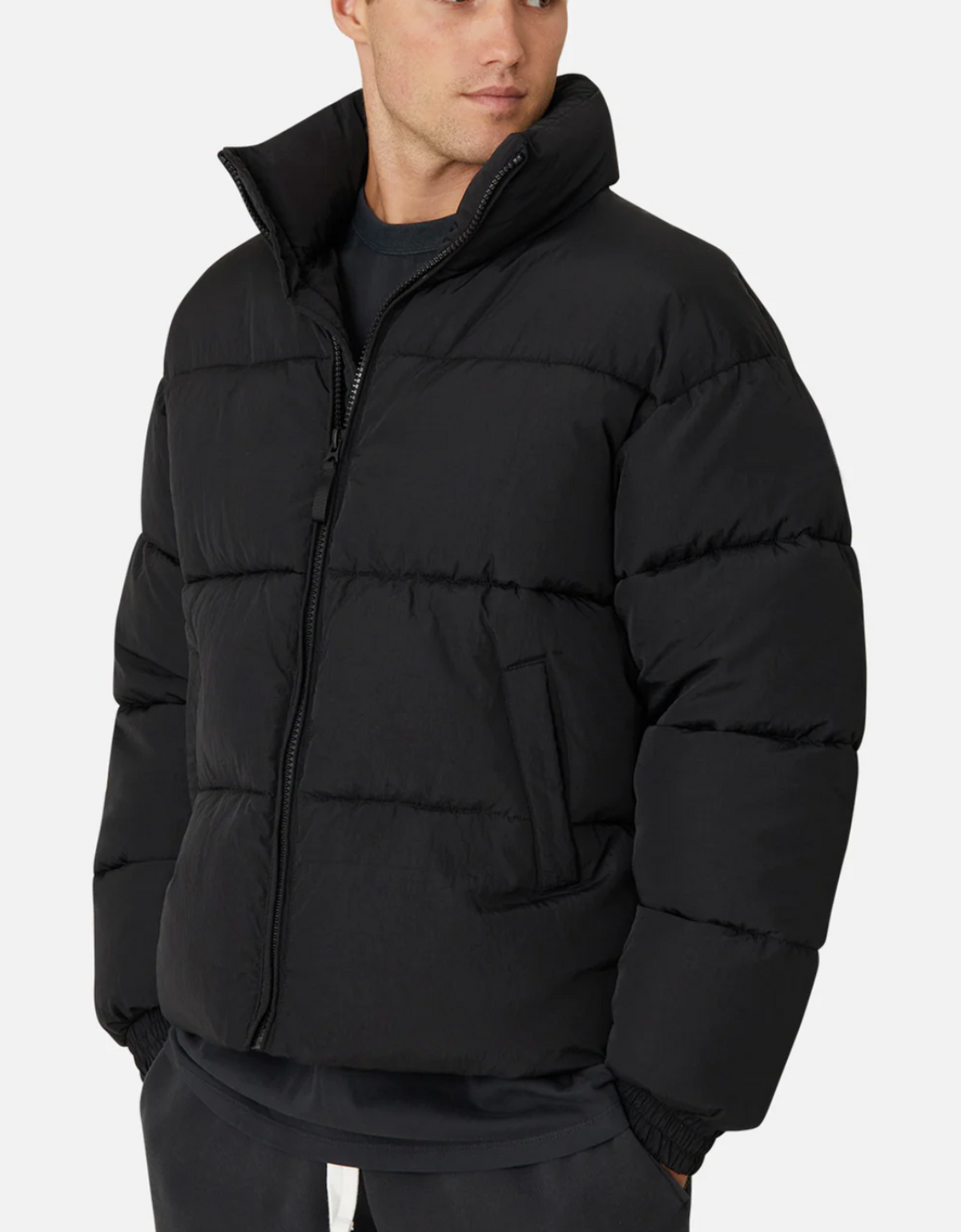 Wearing Industrie The Regent Puffer Jacket—sleek black with quilted padding, high collar, and zipped up—they glance left in dark pants and a shirt. A plain white background highlights this chic ensemble, perfect for an Industrie-inspired look.