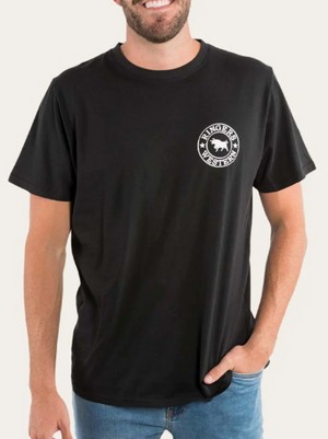 A person in a black Ringers Western Signature Bull Loose Fit T-Shirt, made from 100% cotton jersey with a white circular logo on the left chest, and blue jeans stands smiling with one hand in their pocket.