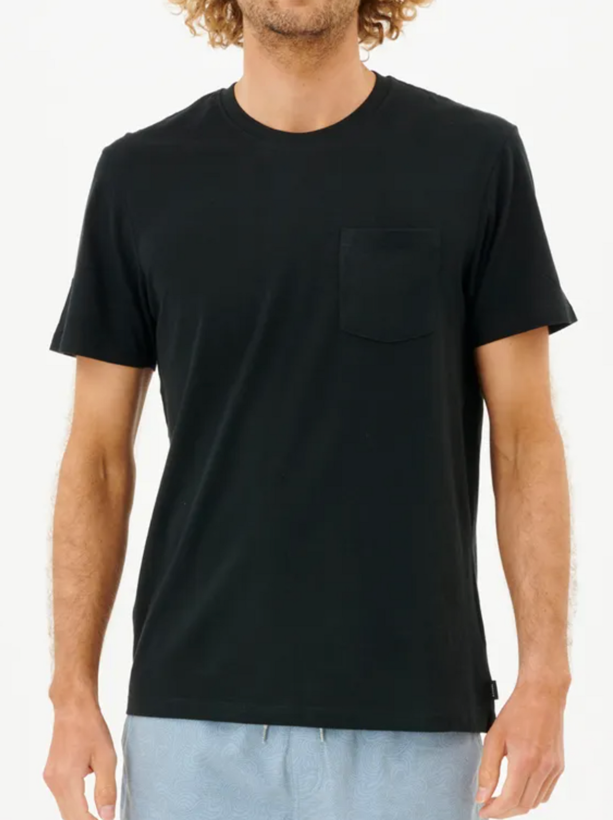 A person stands against a plain background wearing a Rip Curl Plain Pocket Tee in black, made of 100% cotton, paired with light blue shorts. The face is not visible.