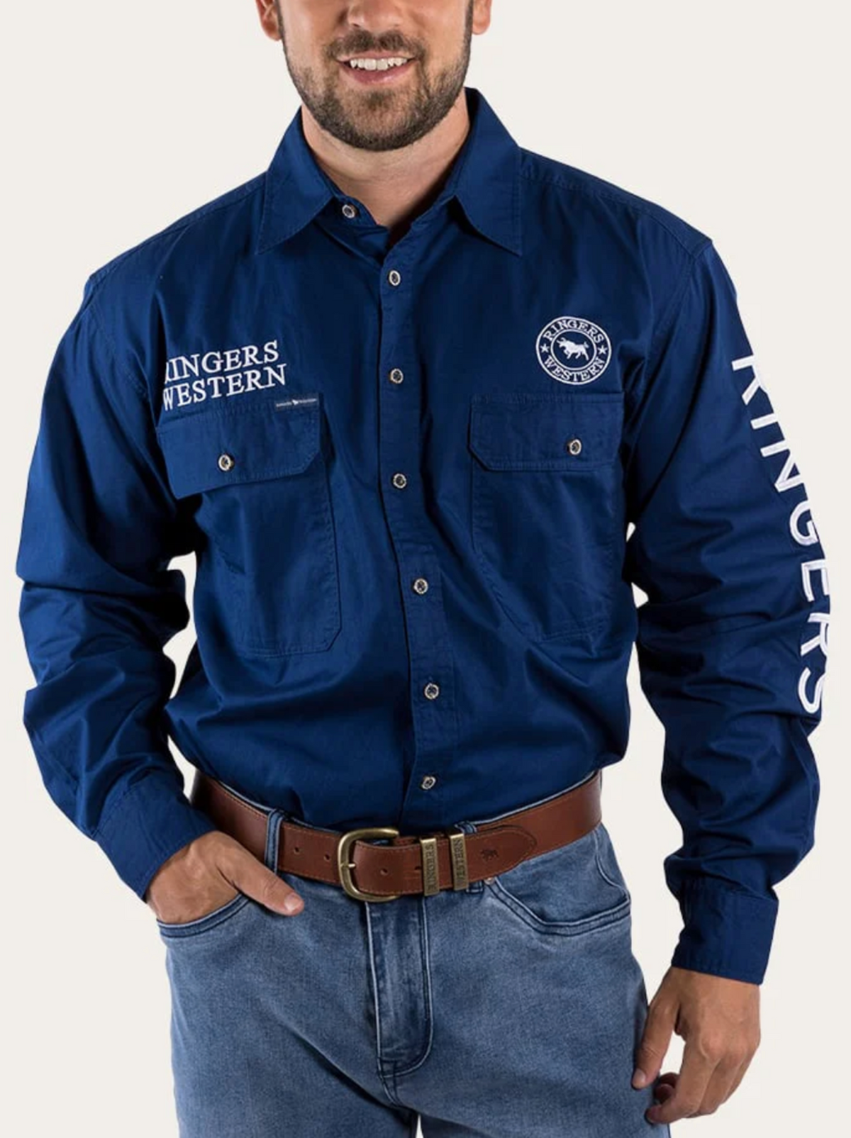 A man wears the Ringers Western Hawkeye Embroidered Shirt in blue, featuring UPF50+ protection, "RINGERS WESTERN" embroidery on the chest and sleeve, two front pockets, a chest logo, paired with light blue jeans and a brown belt.