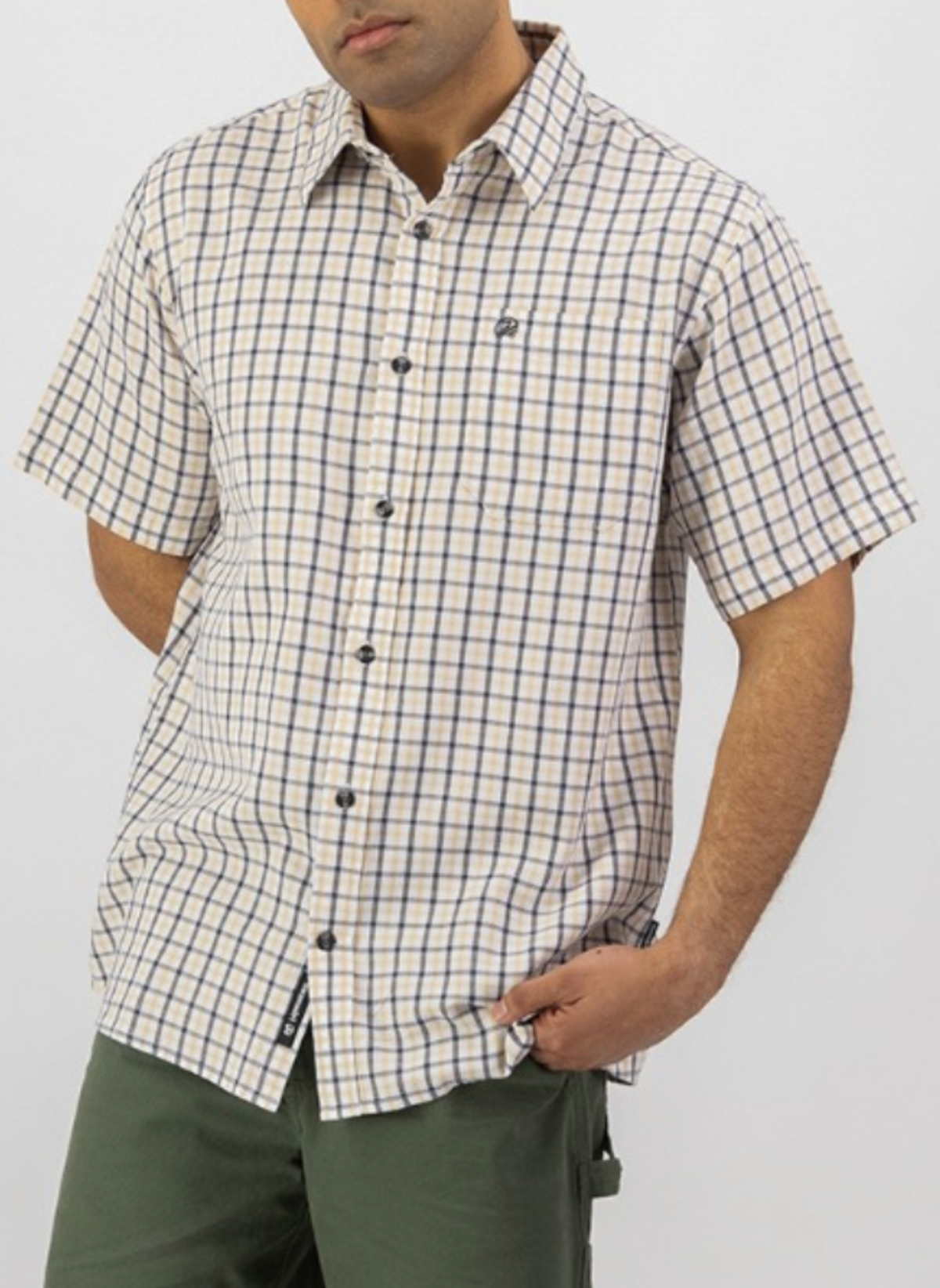 A person wearing the Swanndri Russell Shirt, a relaxed fit, short-sleeve 100% cotton button-up in beige and navy check, and green pants stands with one hand in their pocket against a plain white background.