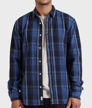 A person is wearing the stylish Academy Brand Douglas Shirt— a blue and black plaid button-up—over a white t-shirt. Made from 100% cotton, the shirt features long sleeves and several white buttons down the front. With its relaxed fit, only the torso and part of the neck are visible against a plain, light-colored background.