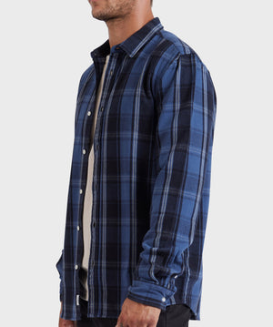 A person wearing an Academy Brand Douglas Shirt, a relaxed-fit blue and black plaid flannel, over a white undershirt. The sleeves of the 100% cotton flannel are buttoned at the cuffs. The individual stands sideways, showing the left profile of their upper body against a plain background.