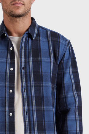 A person wearing the Academy Brand Douglas Shirt, a blue and black plaid button-up overshirt, layered over a white T-shirt. This stylish garment, crafted from 100% cotton and designed with a relaxed fit, showcases its quality. The image captures the upper half of the person's chest and shoulders, including the lower half of their face against a plain white background.