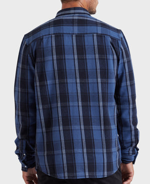 A person with short hair is seen from the back wearing the Academy Brand Douglas Shirt, a stylish long-sleeve overshirt featuring a blue and black checkered pattern. The shirt includes a yoke detail on the shoulders and is made from 100% cotton, offering a relaxed fit. The individual is standing against a plain gray background.