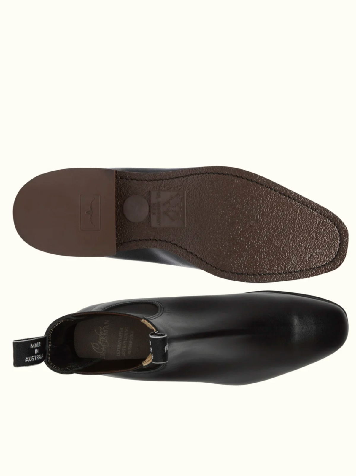 A pair of RM Williams Comfort Craftsman Boots in black is shown. One boot is positioned to display the top view and side profile, emphasizing the smooth pull-up leather surface and elastic side panel. The other boot showcases the durable rubber sole, featuring a textured brown surface with branding, ideal for modern comfort.