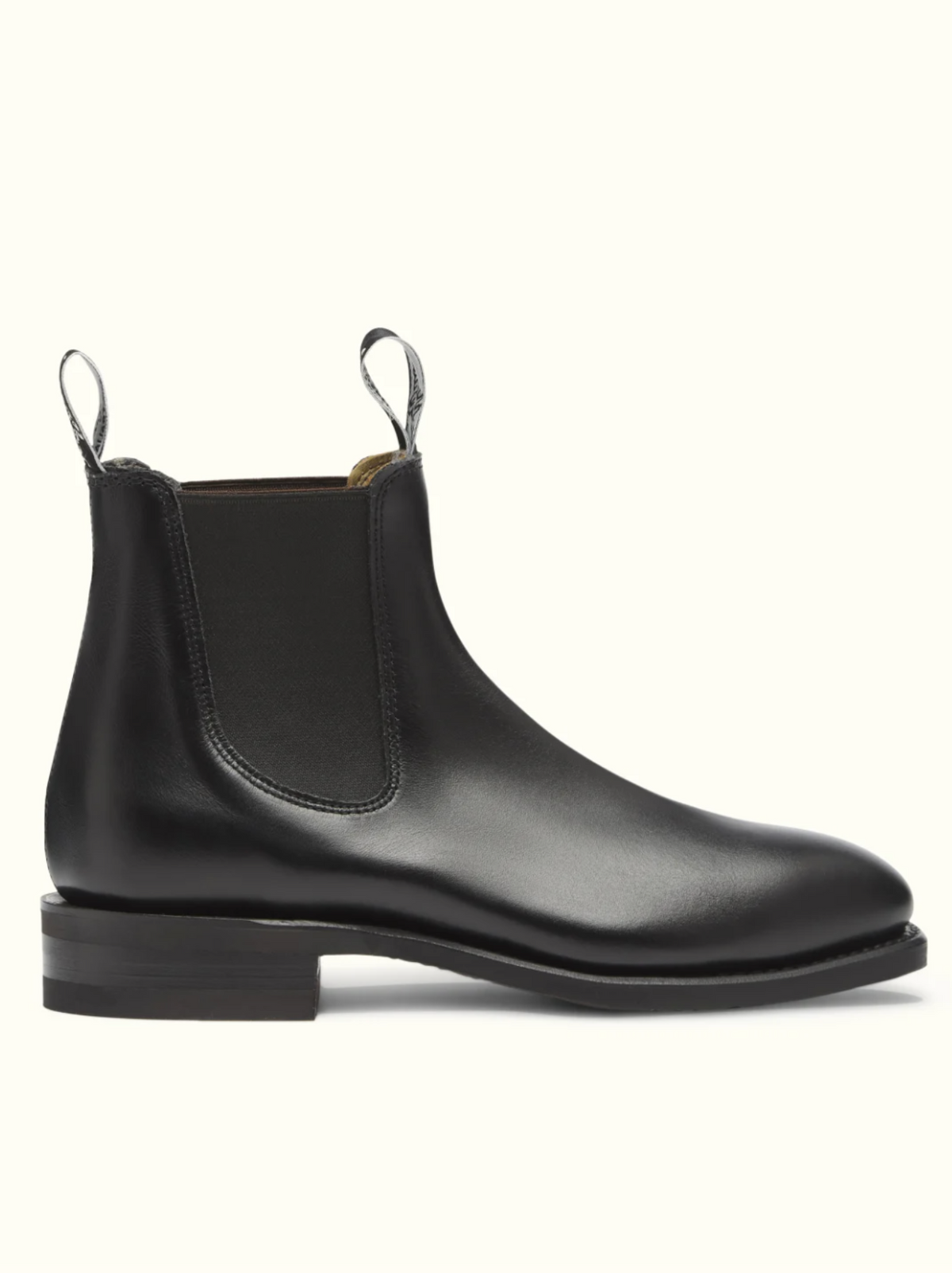 The RM Williams Comfort Craftsman Boot is presented on a plain light background, showcasing its black leather Chelsea design with a smooth finish. It features durable rubber soles, elastic side panels, pull tabs at both the front and back, and a sleek, low wooden heel.