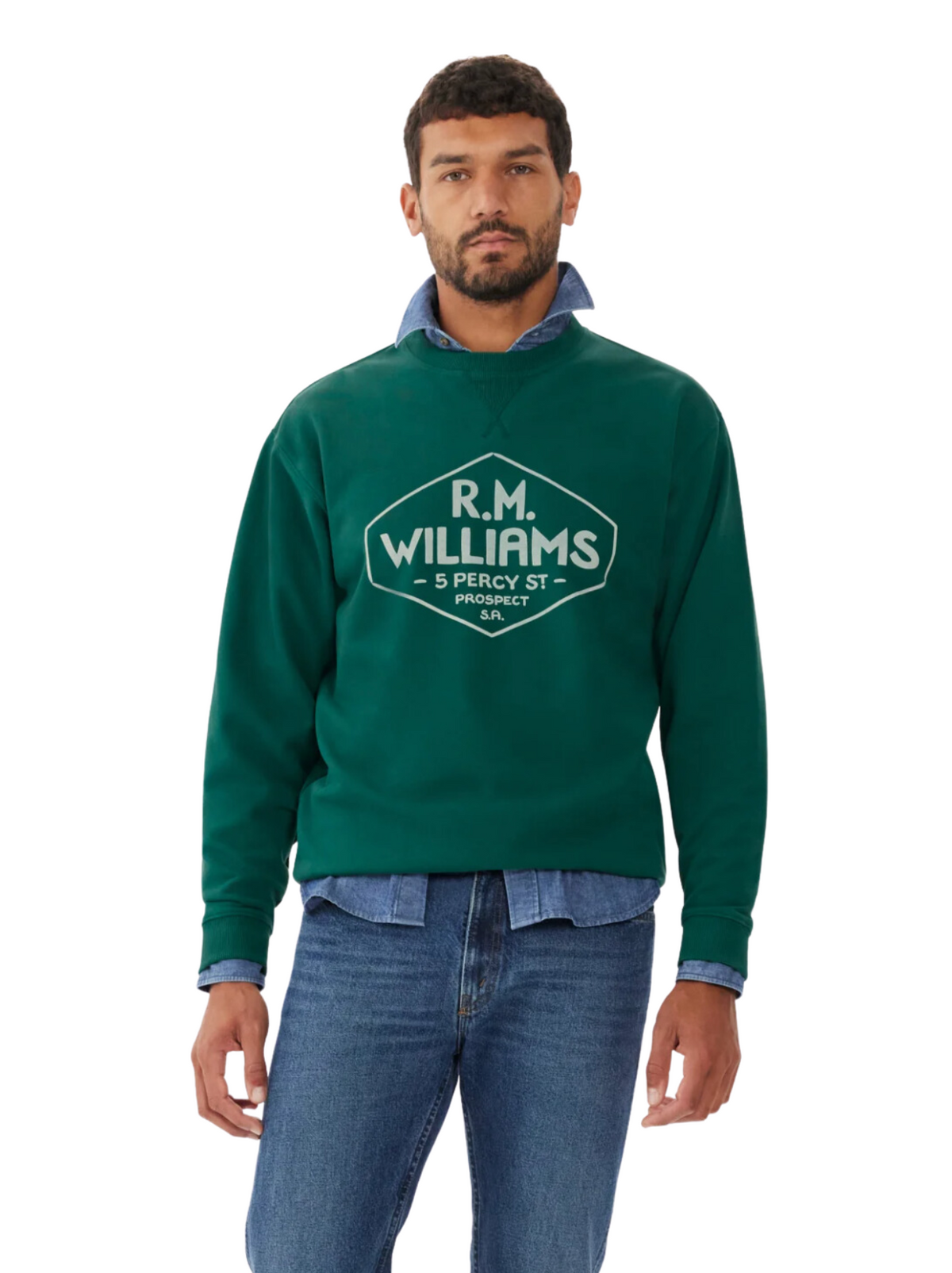A man with short dark hair and a beard wears an RM Williams Gladstone Crew Neck in green cotton fleece, styled over a blue button-up shirt and blue jeans, standing against a plain white background.