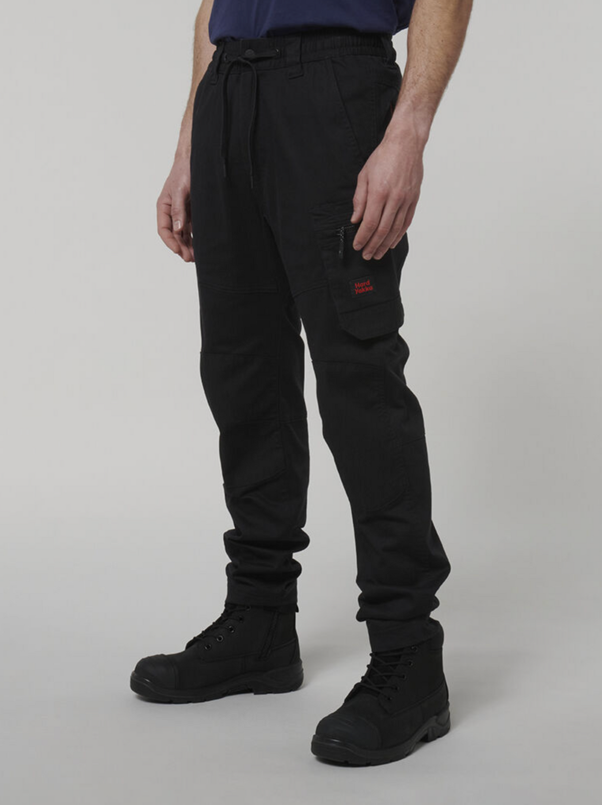 A person wearing black Hard Yakka Tough Maxx Pant cargo pants with UPF 50+ sun protection and black lace-up boots stands against a plain light gray background. Their face is not visible, and their hands are relaxed at their sides.