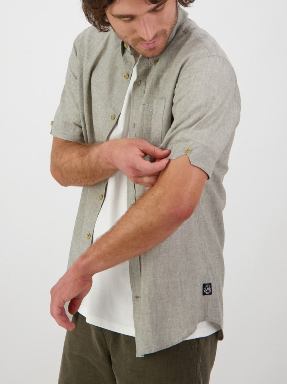 Wearing a Swanndri Lowell v2 Short Sleeve Shirt in light gray, made from a linen-cotton blend, the individual rolls up one sleeve to reveal a white undershirt while standing against a plain white background.