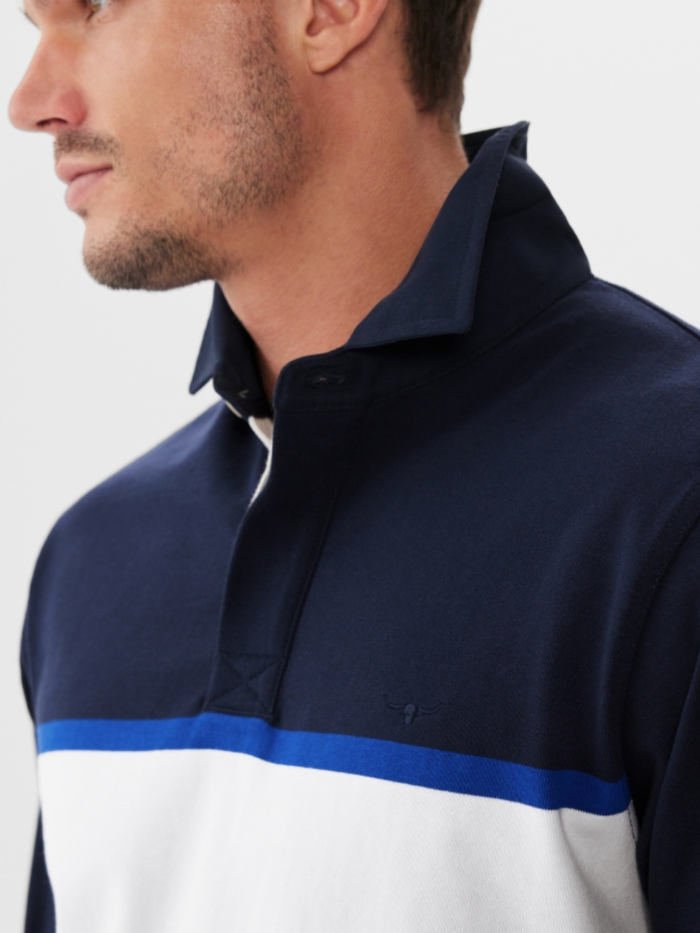 A man with short facial hair wears the RM Williams Tweedale Rugby jersey in navy, featuring a striking white and blue horizontal chest stripe, capturing classic sportswear vibes as he looks to the side against a plain background.