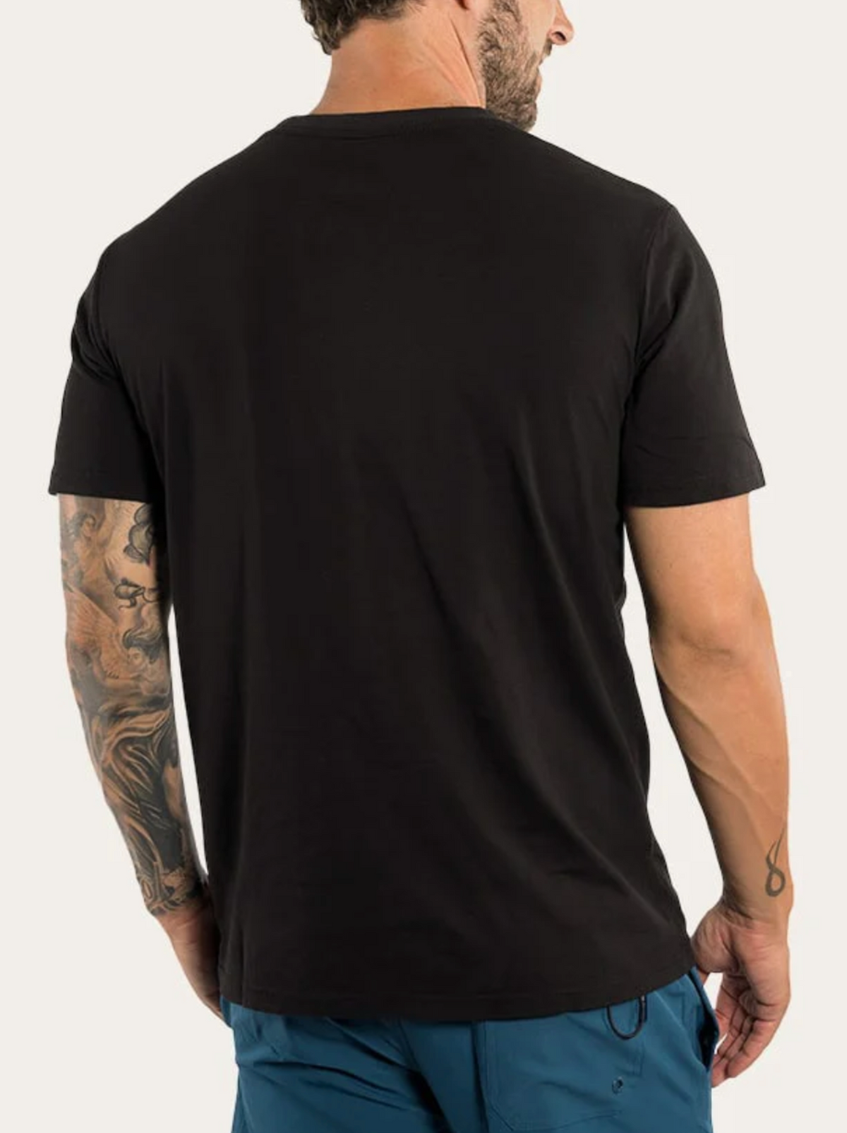 A bearded man with a tattooed arm is shown from the back wearing the Ringers Western The Lodge Classic Fit T Shirt and blue shorts, standing against a plain, light background.